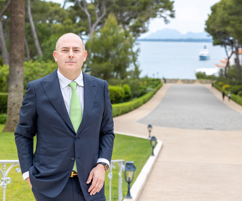Fine Living Services – A picture of Giannis in a suit and green thai with a yacht in the distance behind him